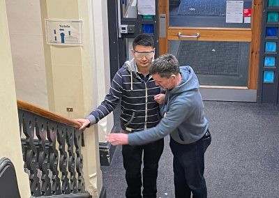 Deafblind workshop - two men guiding, one of them wearing pretence glasses - showing the bannister rail.