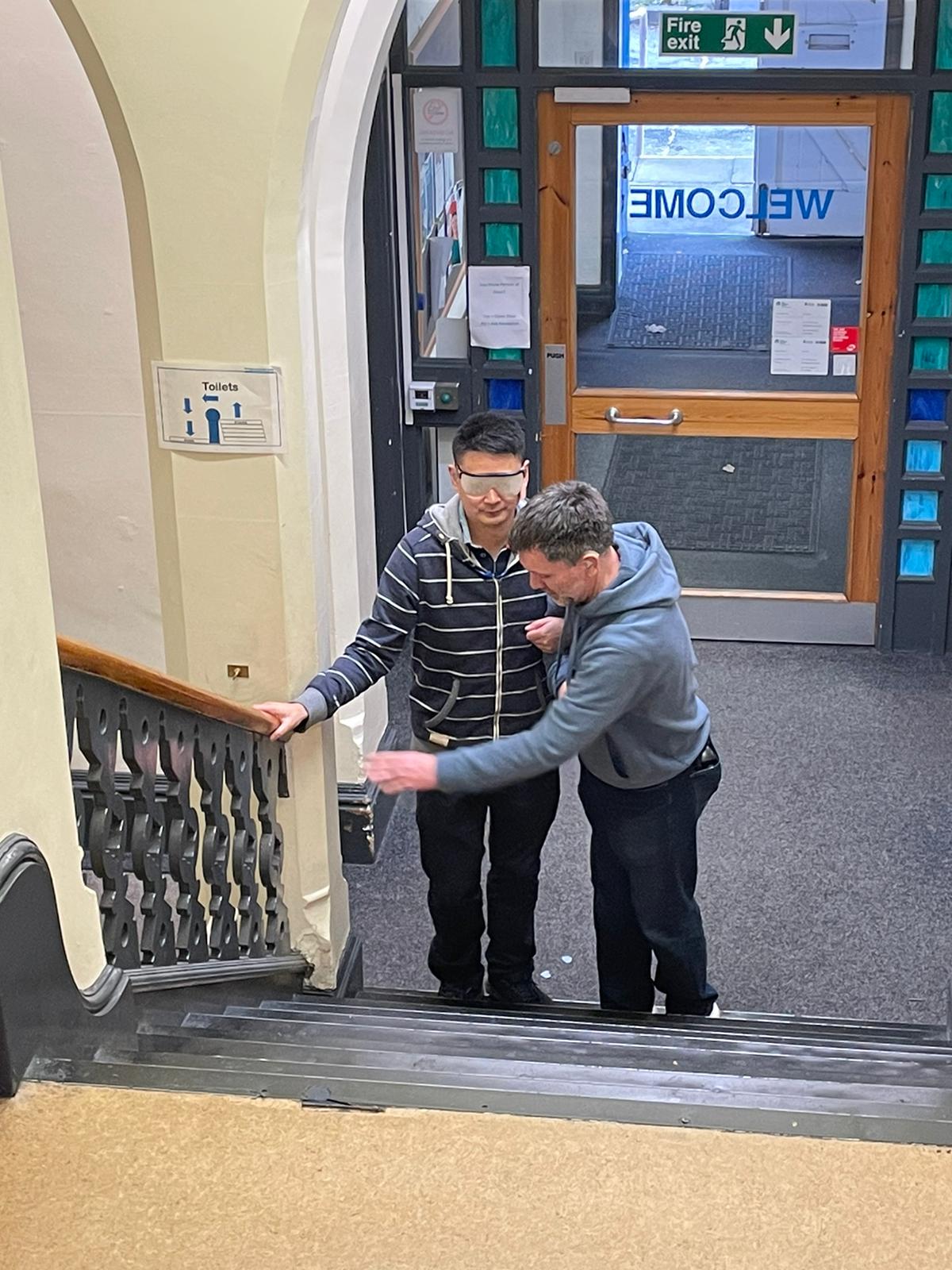 Deafblind guiding taster session - two men at the bottom of the stairs, one is cover with mask and another holding onto him and guiding him up the stair.